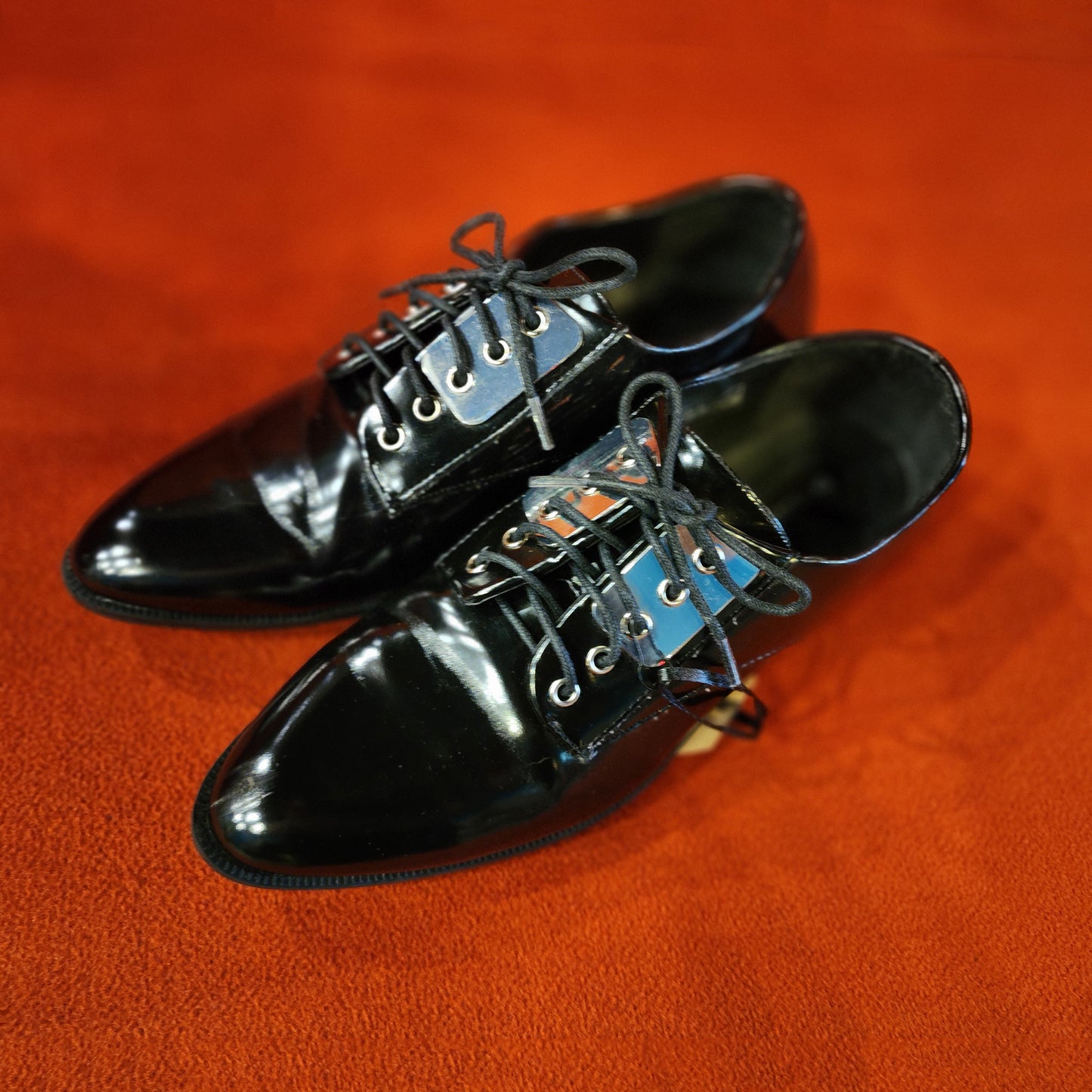 A pair of glossy black leather lace-up dress shoes with silver-toned metal eyelets, resting on a textured orange surface.